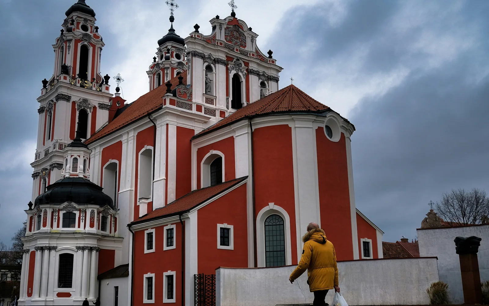 A lone passerby in a yellow coat walks past a stunning baroque church, its red and white façade standing in stark contrast against the moody sky. Standing gracefully in the heart of Vilnius, St. Catherine’s Church (Šv. Kotrynos bažnyčia) is a masterpiece of Baroque architecture. Its striking red-and-white façade, intricate towers, and rich history make it one of the most beautiful landmarks in Lithuania. Despite the overcast sky, the church's elegance remains undeniable, offering a glimpse into Vilnius' vibrant cultural and religious heritage.