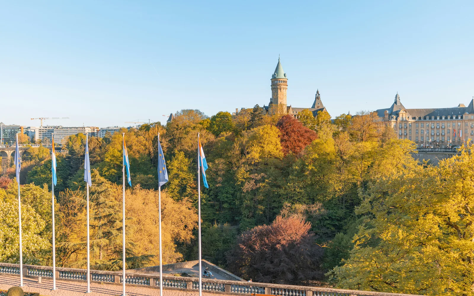 Panoramic view of Luxembourg City with national and European flags, historic buildings, and colorful autumn forest under clear blue sky in Europe.